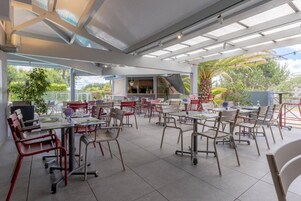 An outdoor dining area with multiple tables and chairs, a covered section with a grill, and a view of the surrounding greenery.