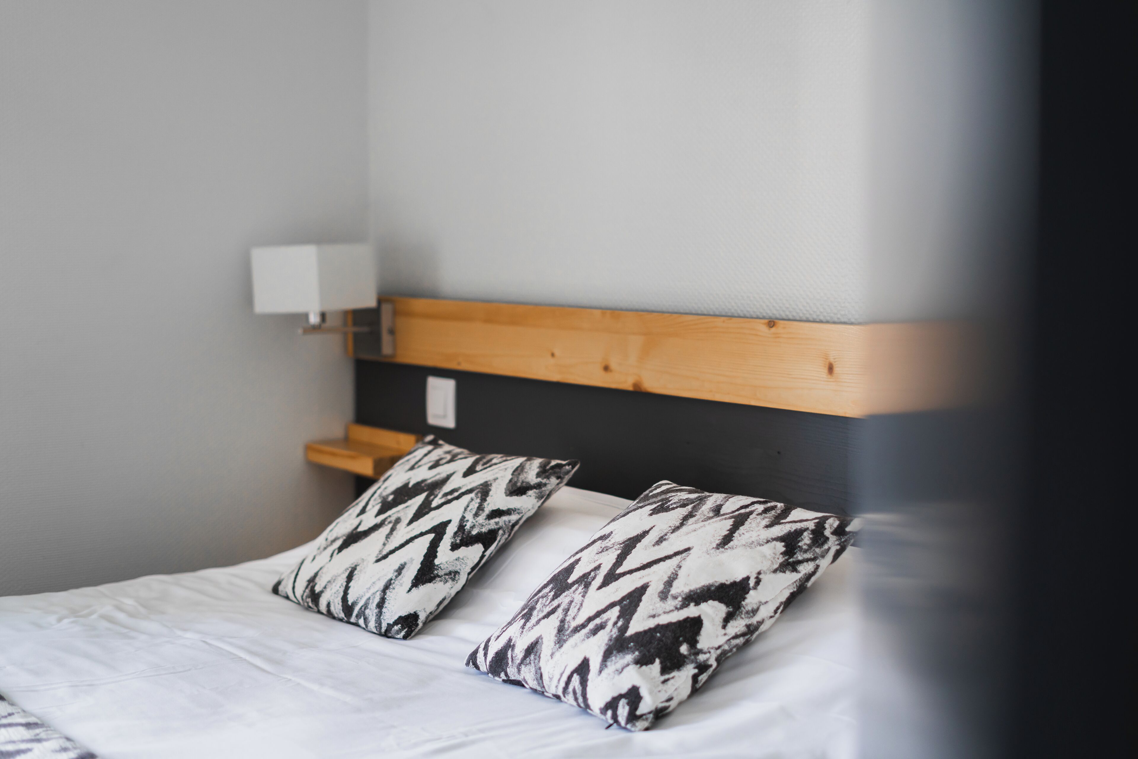 Una cama con dos almohadas estampadas, un cabecero de madera y una mesita de noche blanca con una lámpara.
