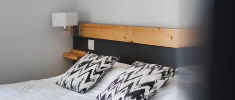 Una cama con dos almohadas estampadas, un cabecero de madera y una mesita de noche blanca con una lámpara.