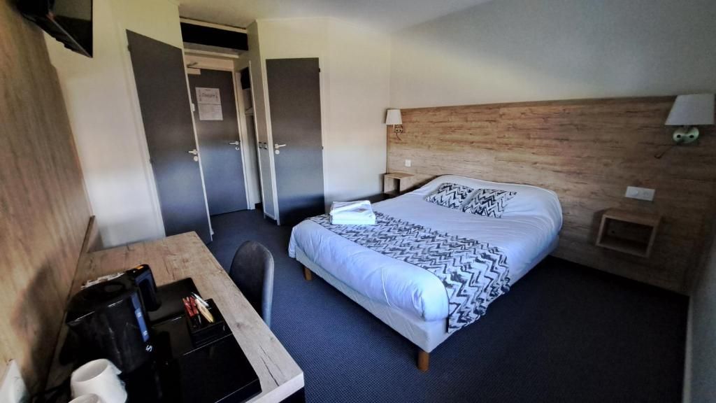 A hotel room with a bed, a desk with a chair, a small table, a nightstand, and a wall-mounted lamp.