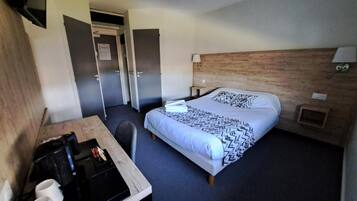 A hotel room with a bed, a desk with a chair, a small table, a nightstand, and a wall-mounted lamp.