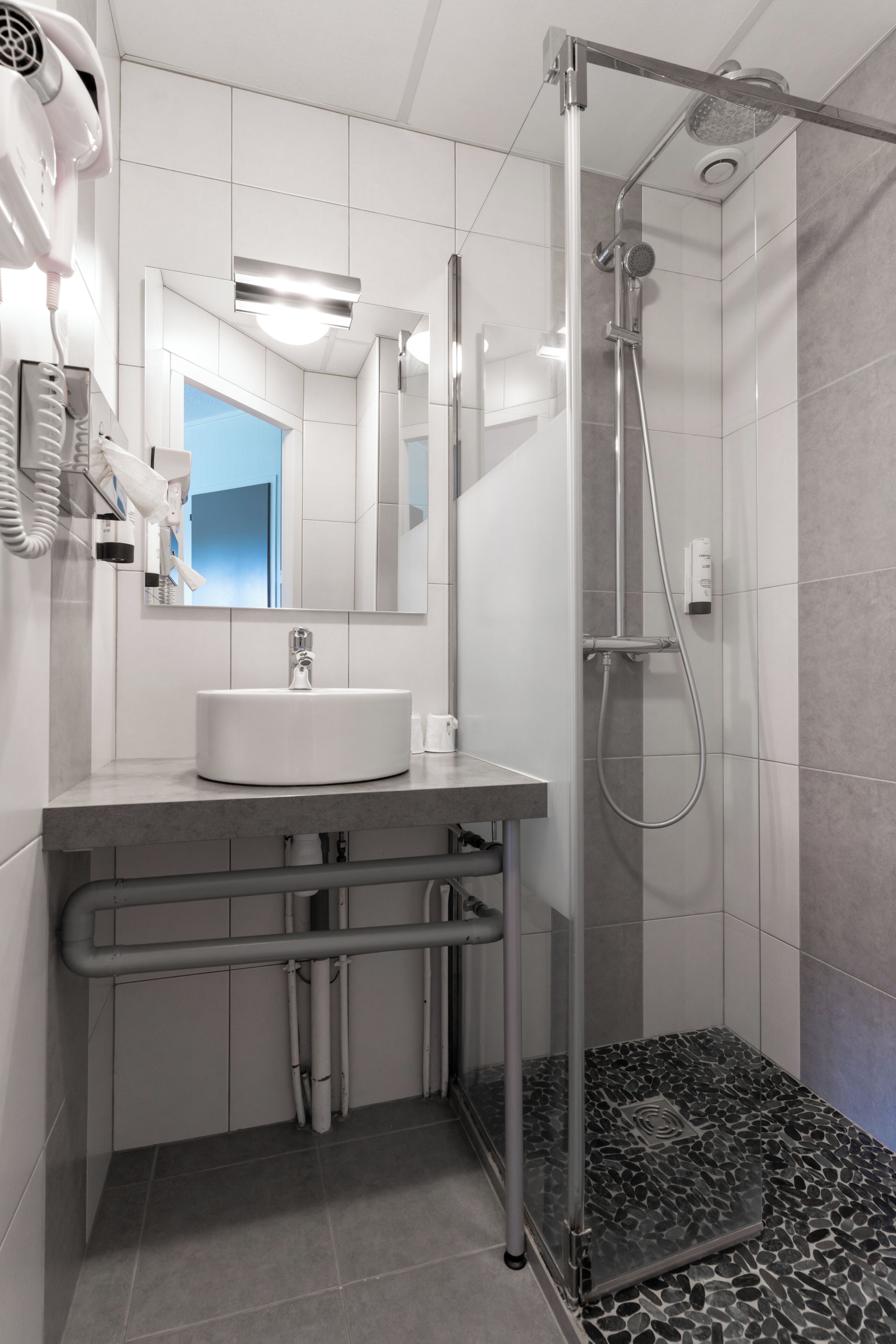 Un baño moderno con lavamanos blanco, cabina de ducha de vidrio y secador de pelo en la pared.