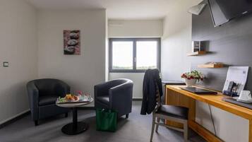Habitació d'hotel amb dues butaques, una taula petita amb un plat de menjar, un escriptori amb un ordinador, un televisor fixat a la paret i una finestra amb vistes a zones verdes.