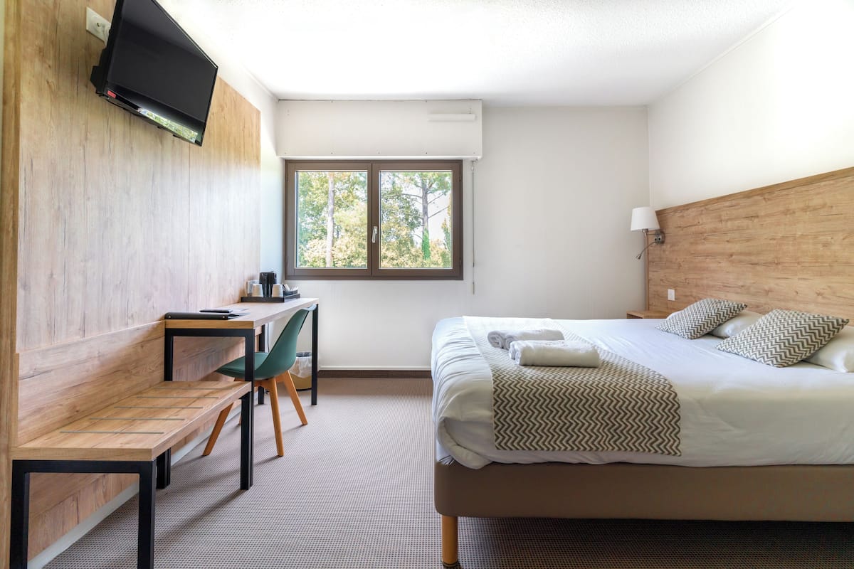 A hotel room with a bed, a desk, a chair, a TV mounted on the wall, and a window with a view of trees.