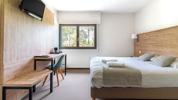 A hotel room with a bed, a desk, a chair, a TV mounted on the wall, and a window with a view of trees.