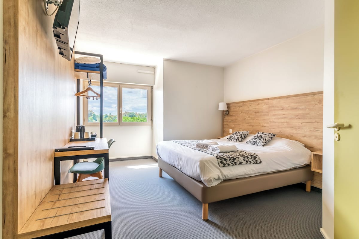 A bedroom with a bed, a desk, a chair, a window, and a wooden headboard.