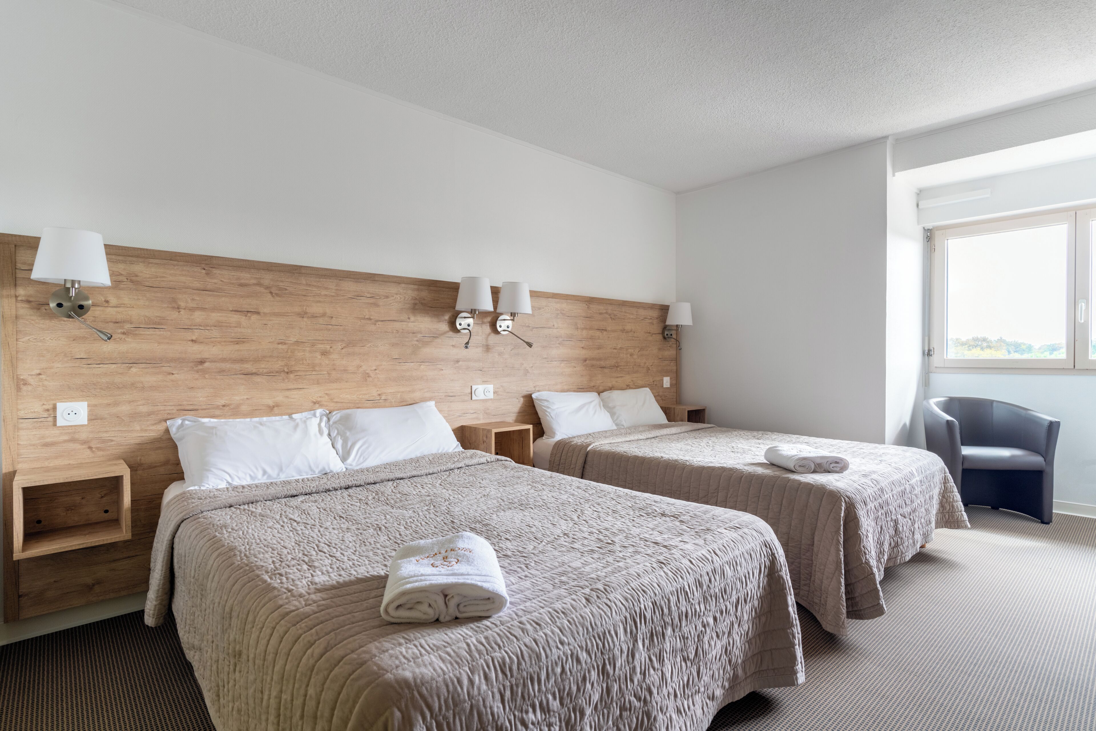 A hotel room with two beds, wooden headboard, bedside lamps, a chair, and a window.