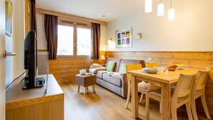 A cozy room with a wooden floor, a sofa, a dining table with chairs, a TV, and a window with curtains. - Residence Pierre & Vacances Les Gemeaux (La Plagne-Tarentaise)