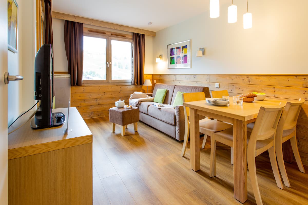 Una habitación acogedora con suelo de madera, un sofá, una mesa de comedor con sillas, un televisor y una ventana con cortinas.