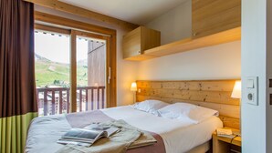 A hotel room with a large bed, wooden headboard, two bedside lamps, a nightstand, and a view of a green landscape through a sliding door. - Residence Pierre & Vacances Les Gemeaux (La Plagne-Tarentaise)