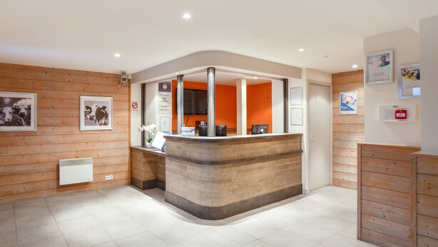 A reception area with a wooden reception desk, tiled floor, and framed pictures on the walls. - Residence Pierre & Vacances Les Gemeaux (La Plagne-Tarentaise)