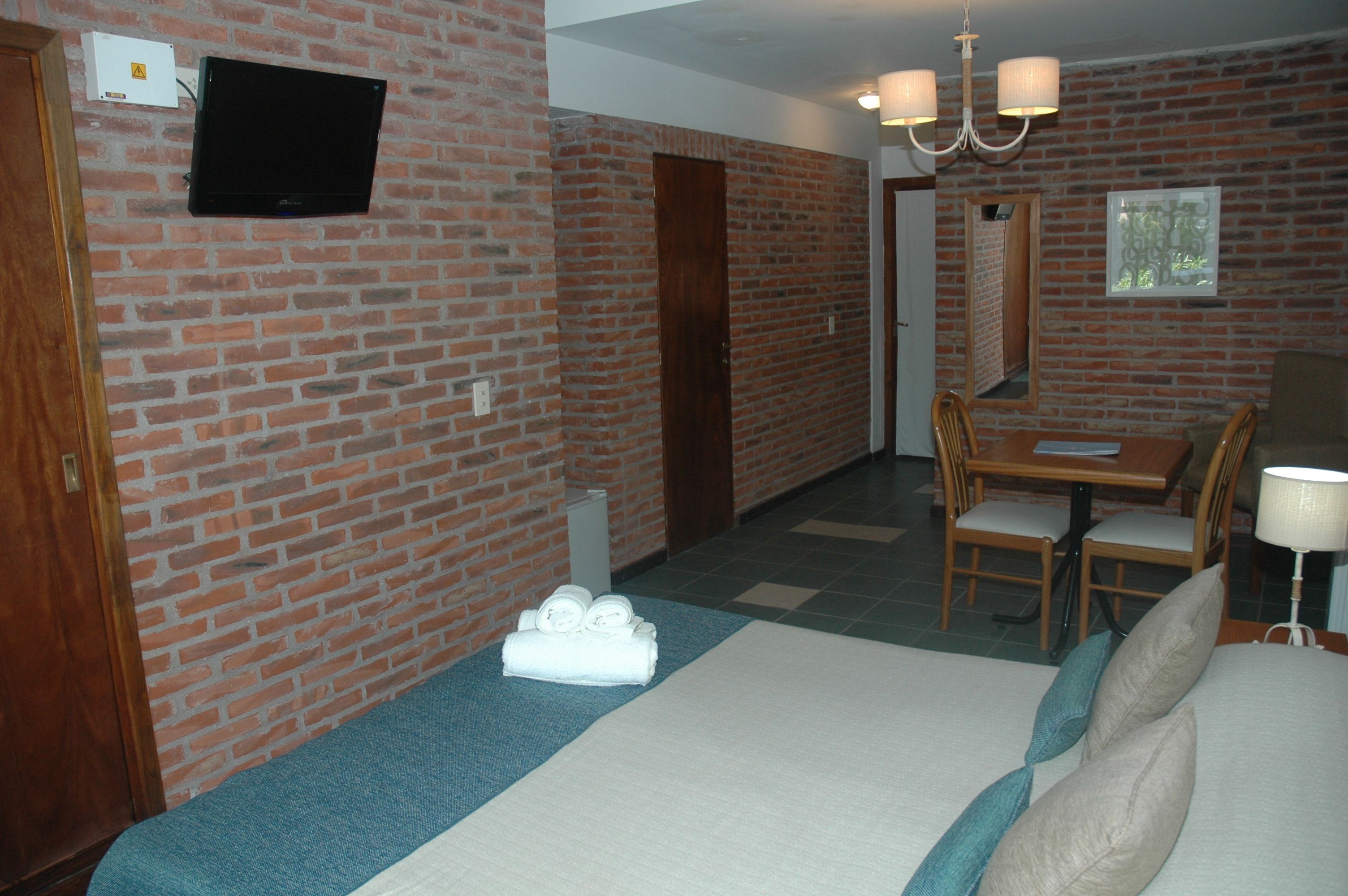 A hotel room with a brick wall, a flat-screen TV, a bed with pillows, a desk with chairs, and a chandelier.