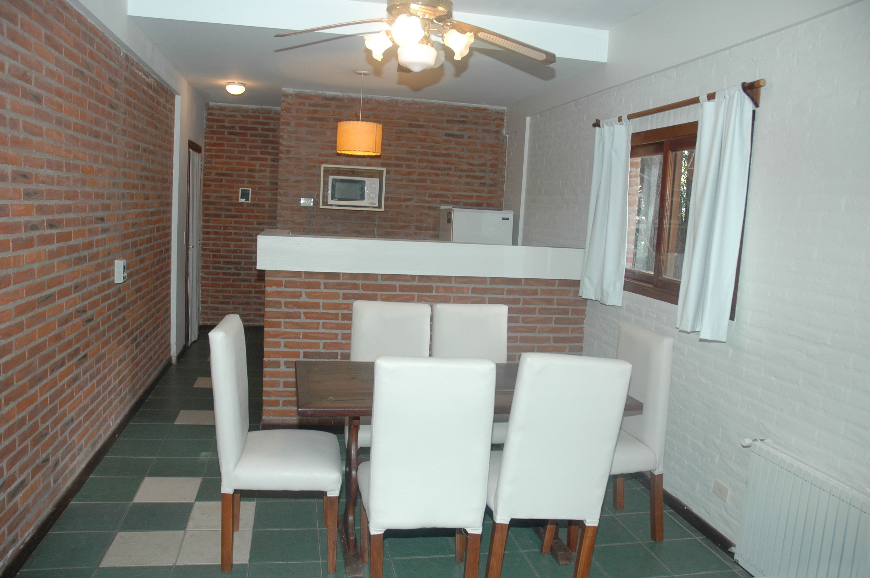 A dining area with a brick wall, white chairs, a wooden table, a ceiling fan, and a window with curtains.