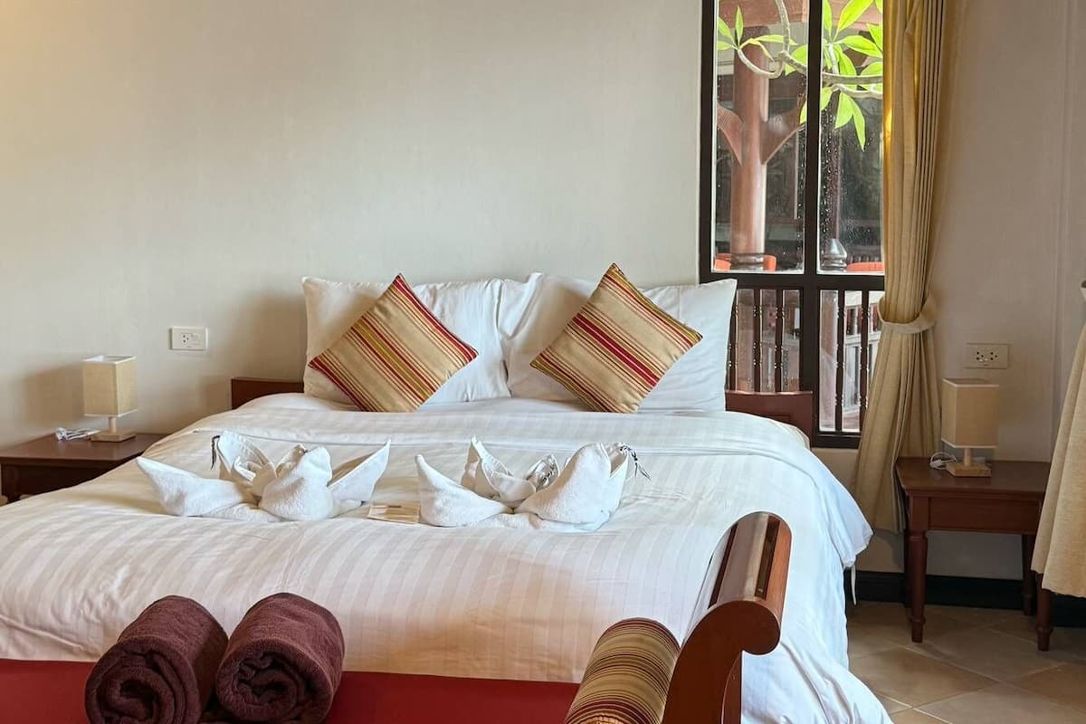 A neatly made bed with white linens and striped pillows, a red bench with towels, and a window with curtains.