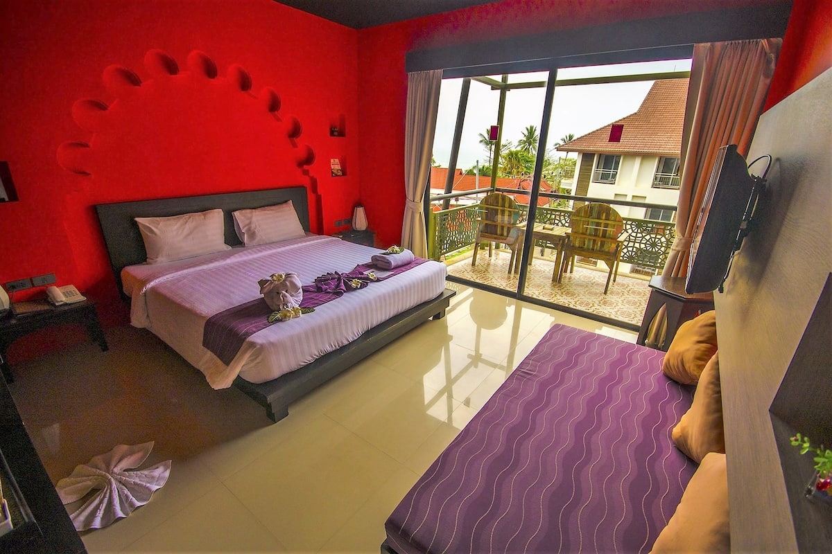 A hotel room with a large bed, a smaller bed, a balcony with a table and chairs, and a view of a building with a red roof.