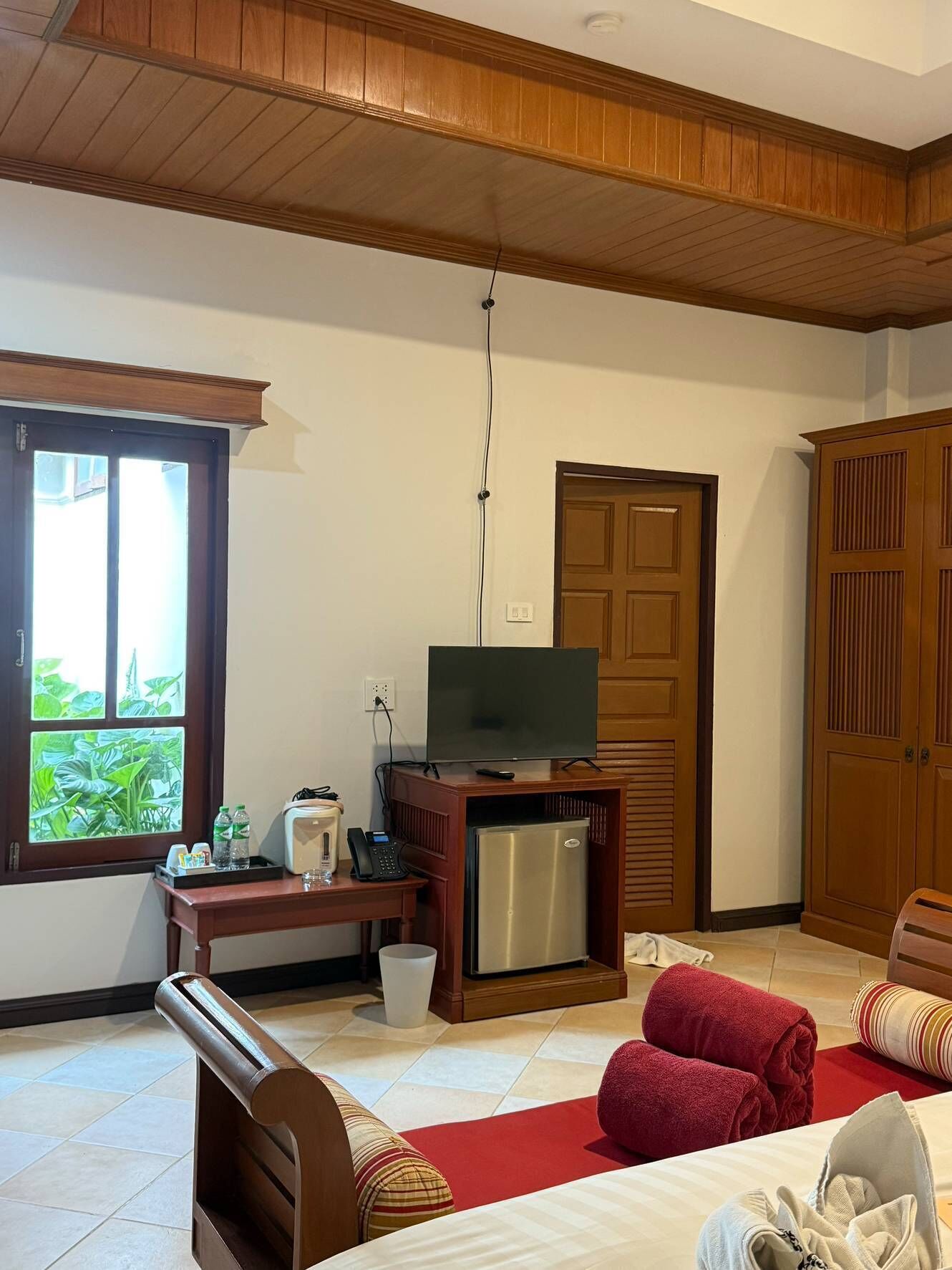A hotel room with a wooden ceiling, a television on a stand, a refrigerator, a desk with a telephone, and a window with a view of greenery.