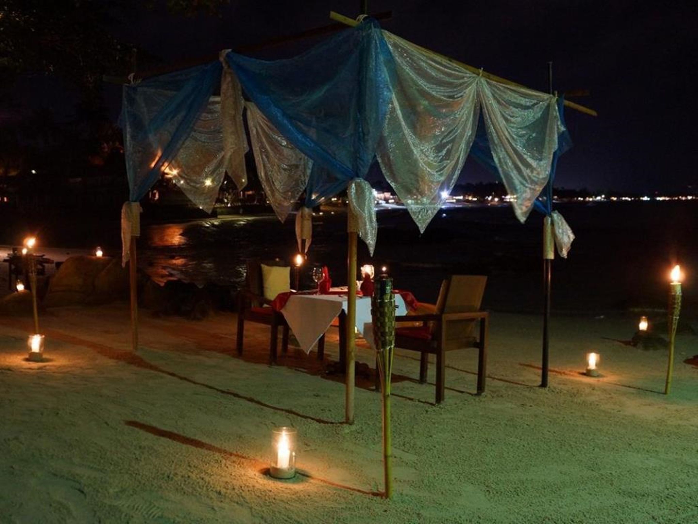 A beachside dining setup with a table and chairs under a blue canopy, surrounded by candles.
