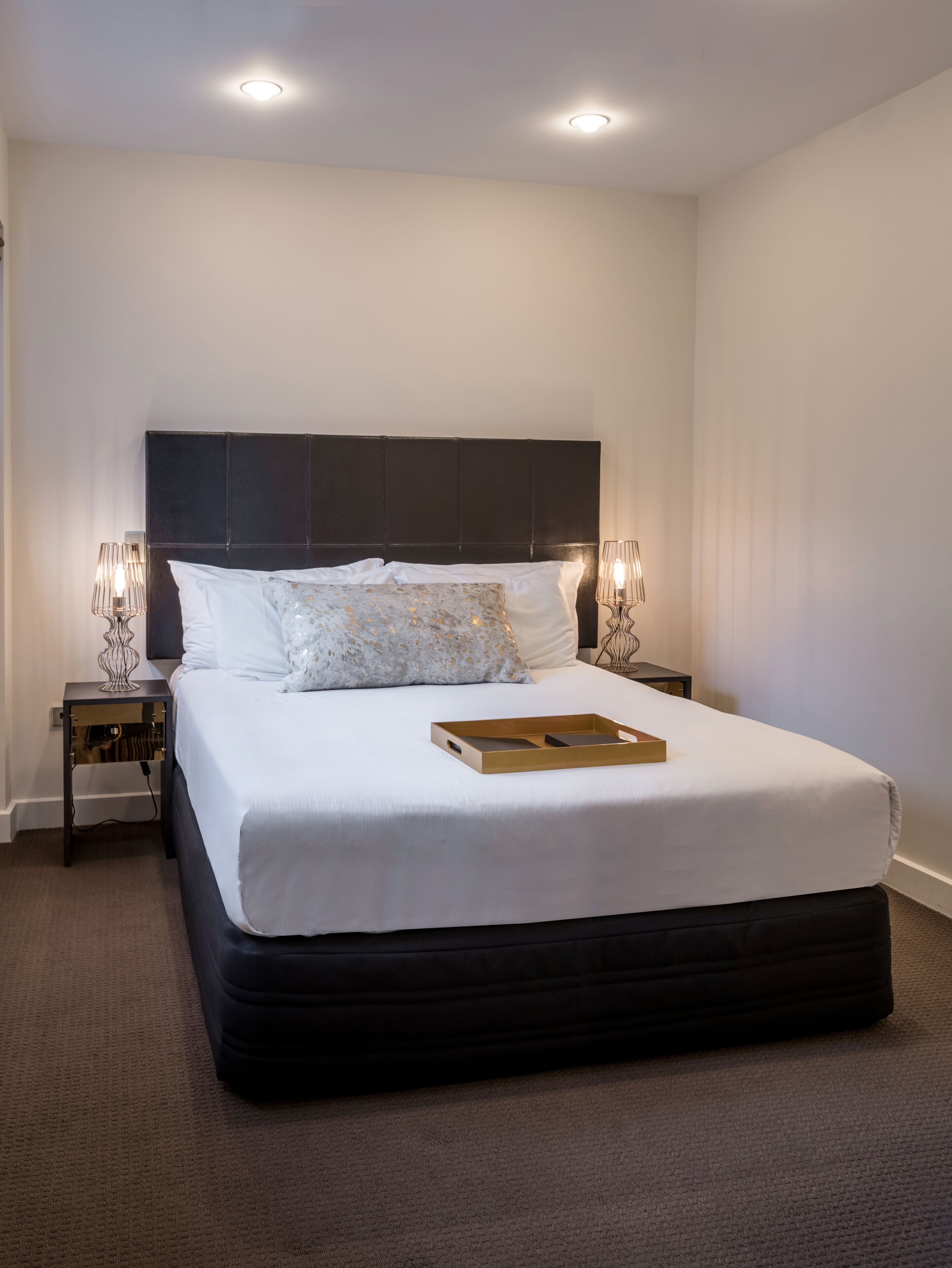 A neatly made bed with a dark headboard, two bedside lamps, and a tray on the bed.