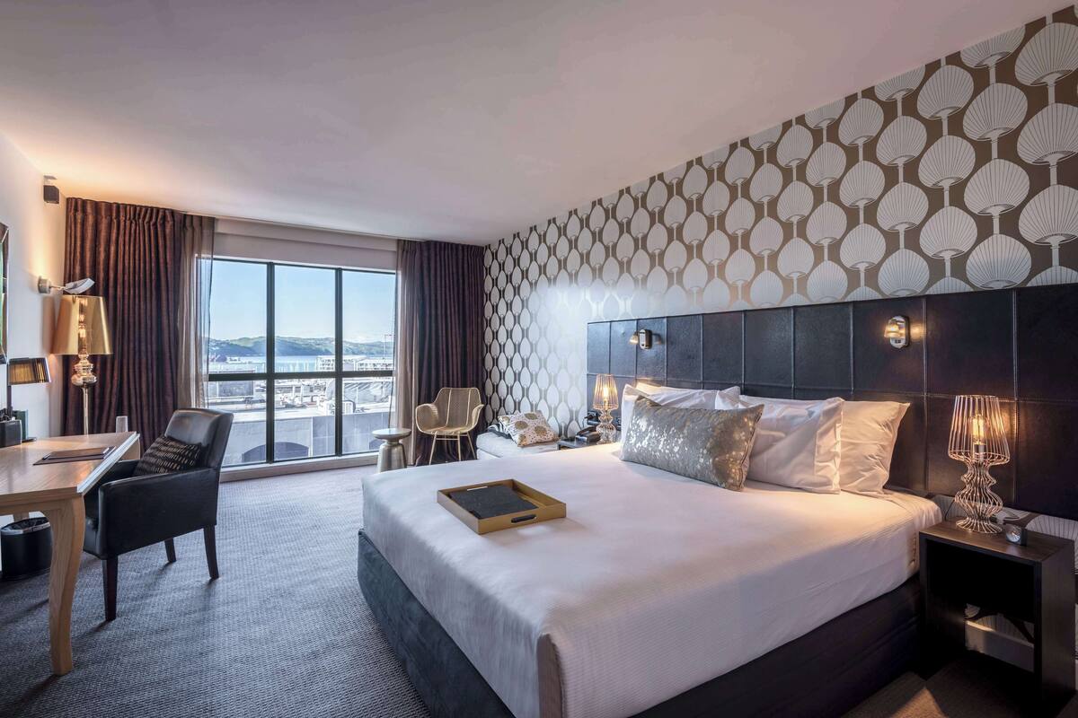 A hotel room with a large bed, a desk, a chair, and a view of the city through large windows.