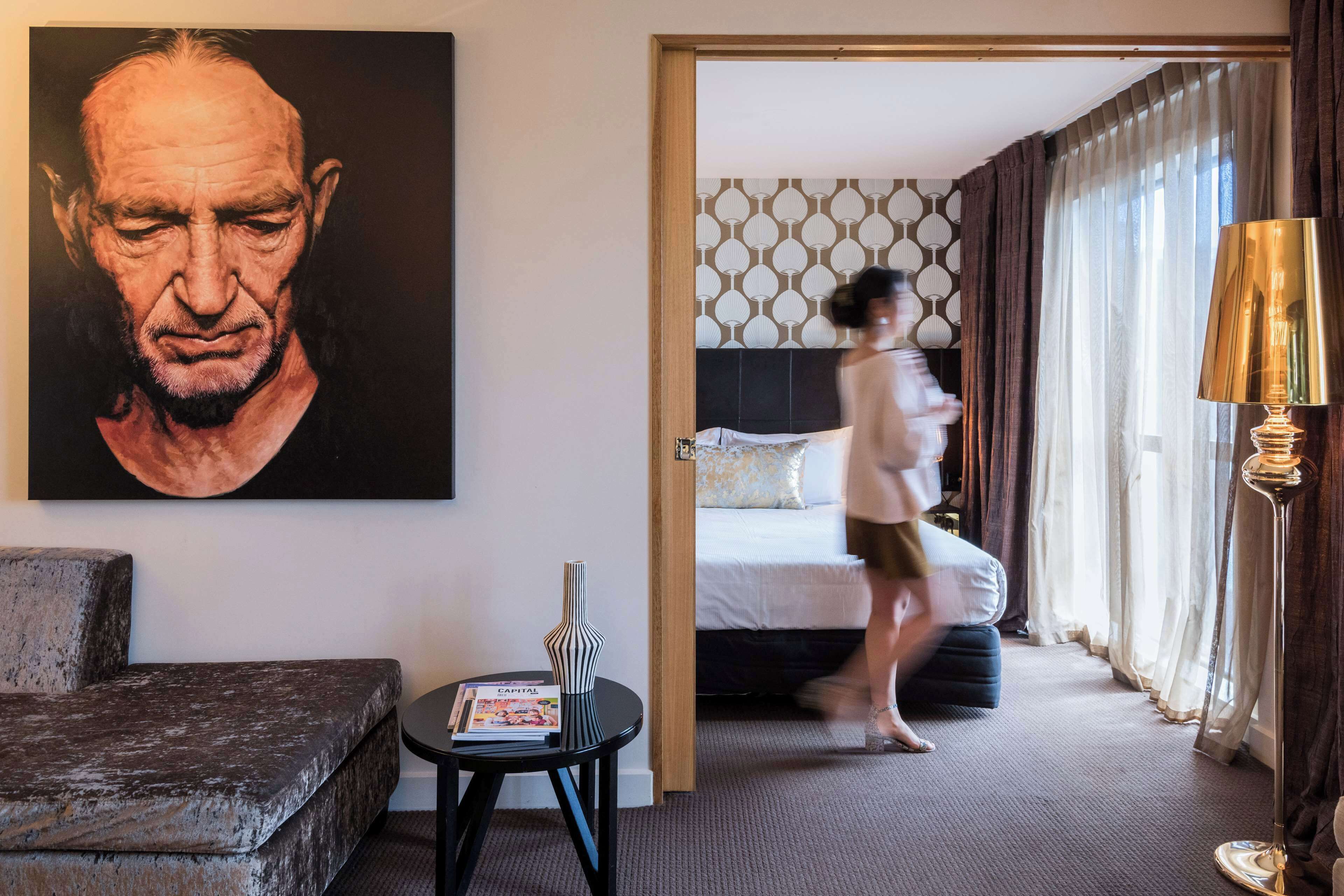 Habitación de hotel con un retrato grande en la pared, una cama con ropa de cama estampada, una mesita de noche con un jarrón y una lámpara de pie.