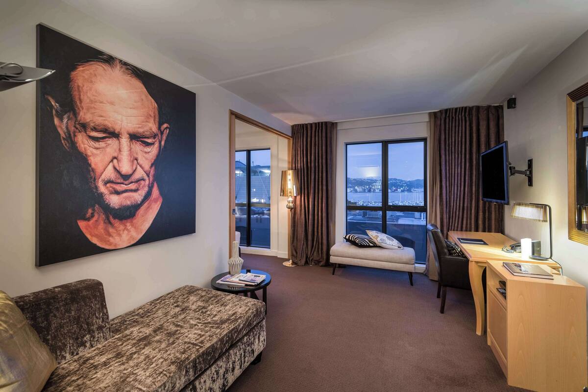 A hotel room with a large painting, a sofa, a desk, a chair, a TV, and a view of the city.