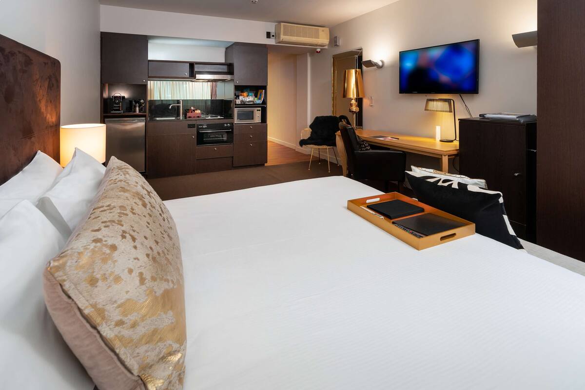 A hotel room with a large bed, a desk with a chair, a television, and a kitchenette.