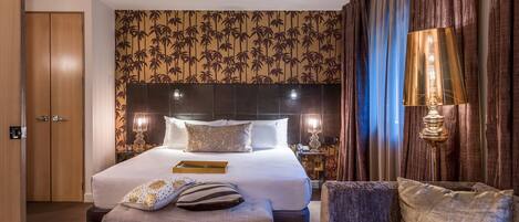 A hotel room with a large bed, a bedside table with a lamp, a wooden wardrobe, and a patterned wall.