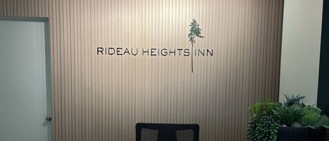 A reception desk at Rideau Heights Inn with a wooden panel wall, a reception area, a chair, a table with a magazine, and a potted plant.