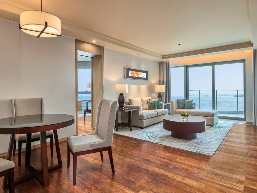 A modern hotel room with a dining area, a sofa, a coffee table, and a large window offering a view of the sea.