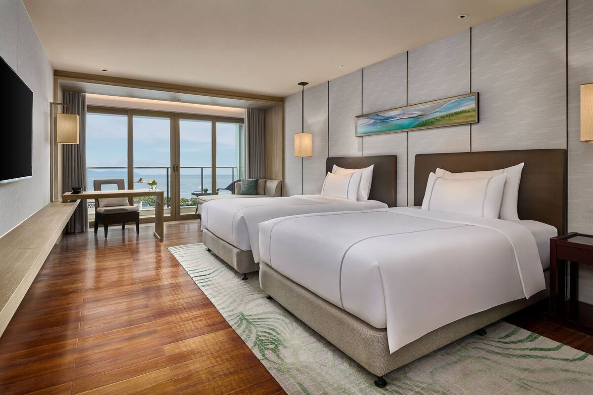 A hotel room with two beds, a desk, a chair, a TV, and a large window with a view.