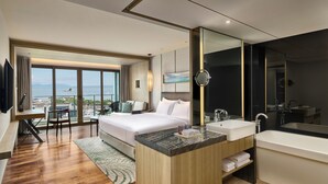 Una habitación de hotel moderna con una cama grande, un espacio de descanso con vistas, un baño con bañera y lavamanos, y un espejo amplio.