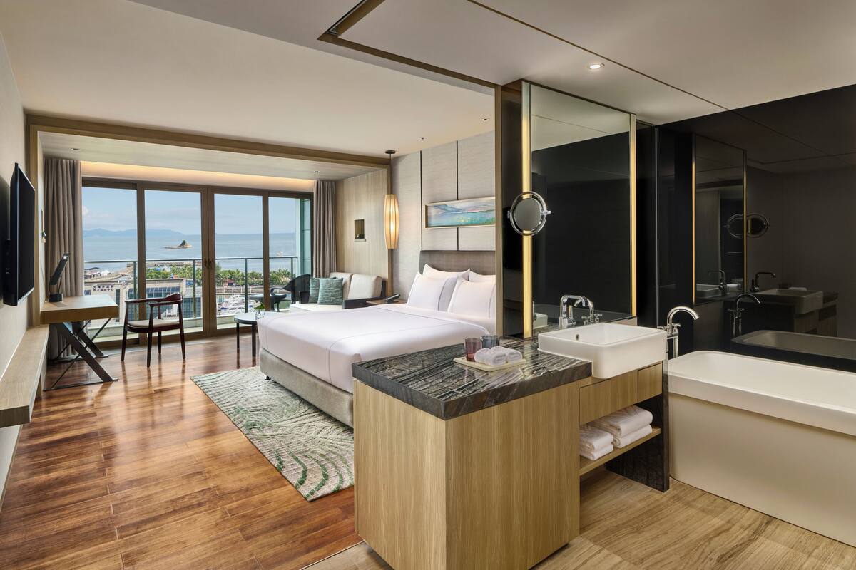 A modern hotel room with a large bed, a sitting area with a view, a bathroom with a bathtub and sink, and a large mirror.