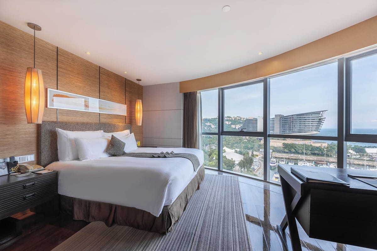 A modern hotel room with a large bed, a desk, and a view of the city through large windows.