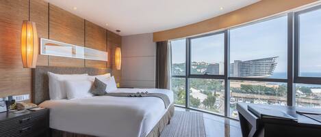 A modern hotel room with a large bed, a desk, and a view of the city through large windows.
