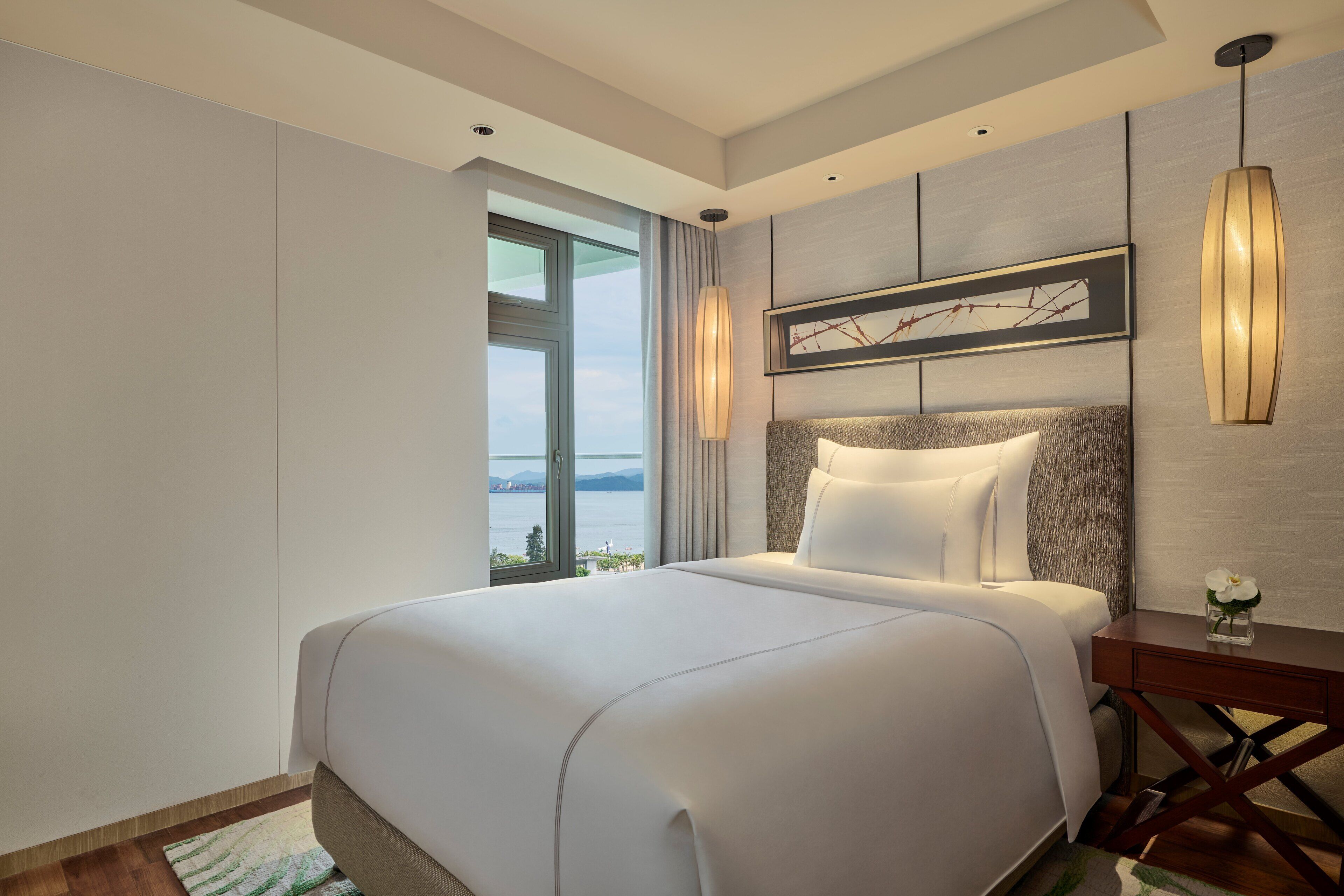 A modern hotel room with a large bed, a view of the outdoors, and a framed artwork on the wall.