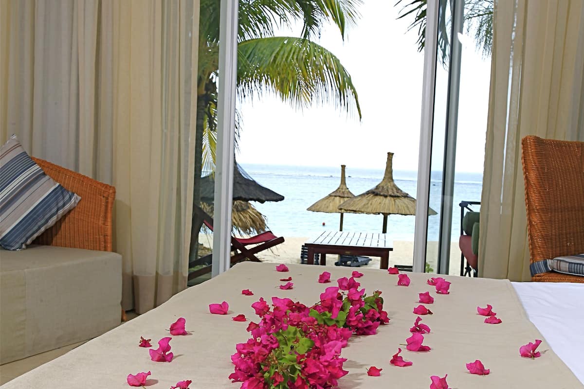 Habitación de hotel con una cama decorada con pétalos de flores rosas, vista a la playa y una palmera.