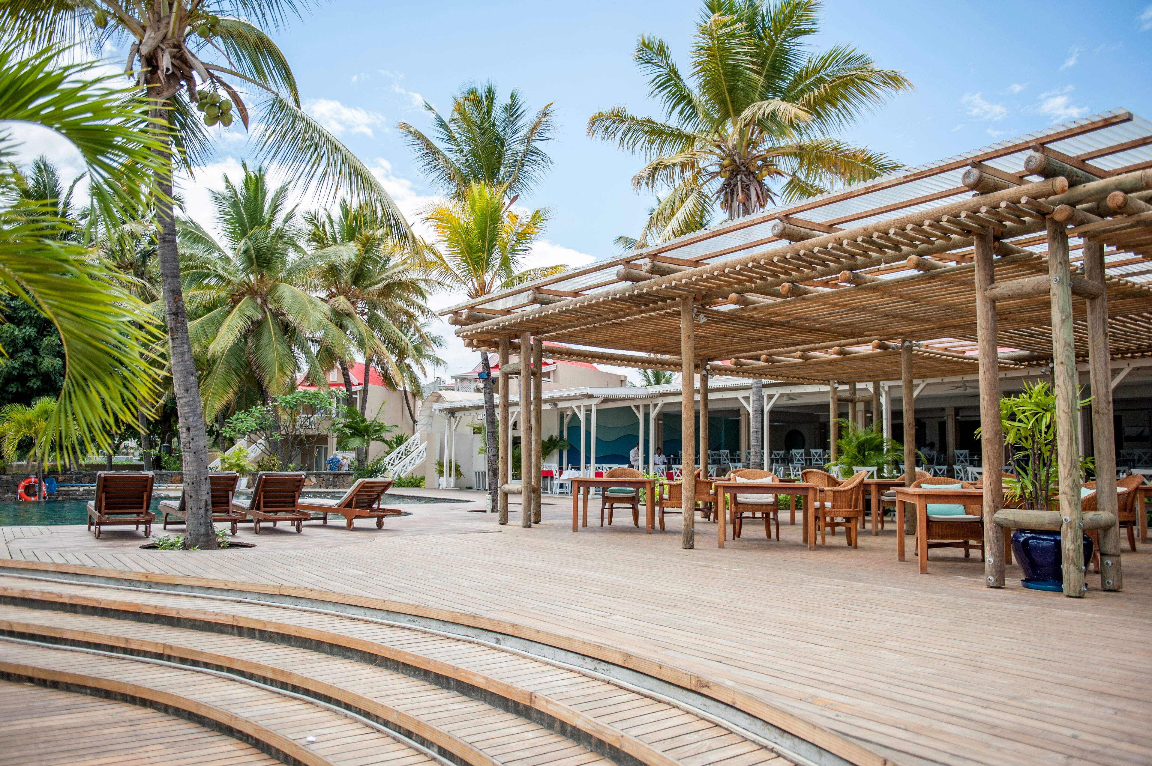 Une terrasse en bois, agrémentée de chaises longues et de tables, entourée de palmiers et bordée d’une piscine.
