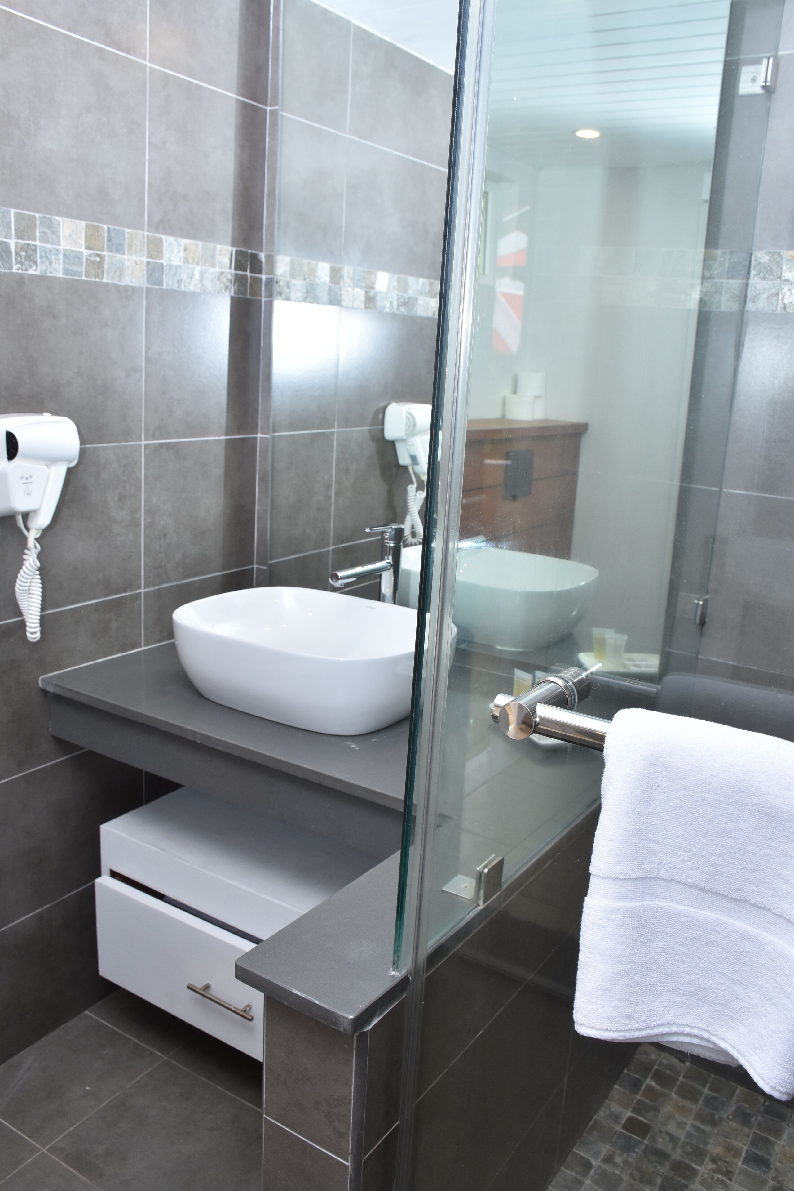 Un baño moderno con ducha de vidrio, lavamanos blanco y secador de pelo en la pared.