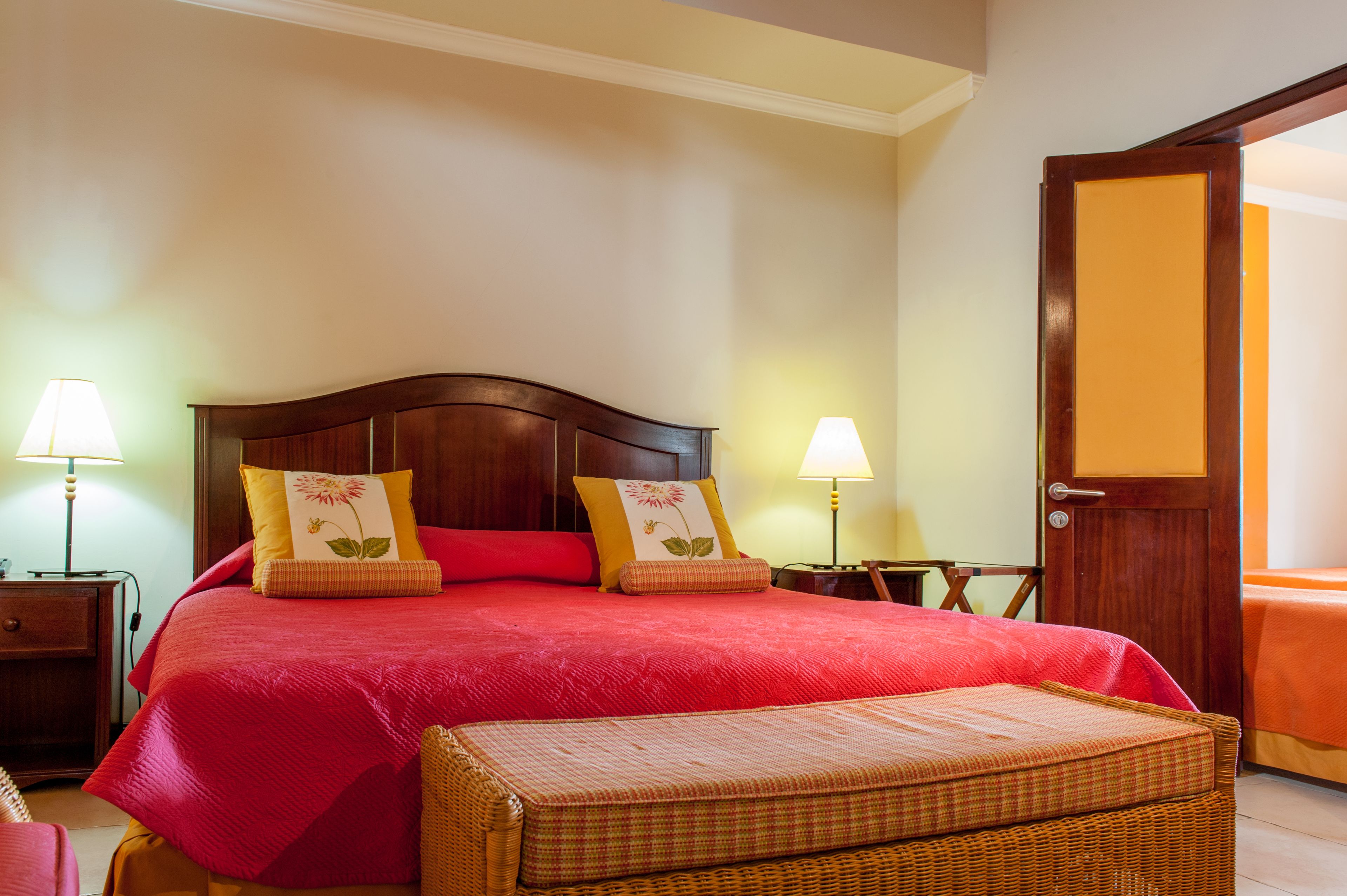 Un letto rifatto con una coperta rossa e due cuscini, una panca in vimini e due abat-jour.