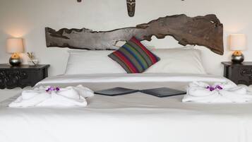 Cama bem arrumada com roupa de cama branca, almofada listrada colorida e cabeceira de madeira. Há duas mesas de cabeceira com abajures e um objeto decorativo na parede acima da cama.