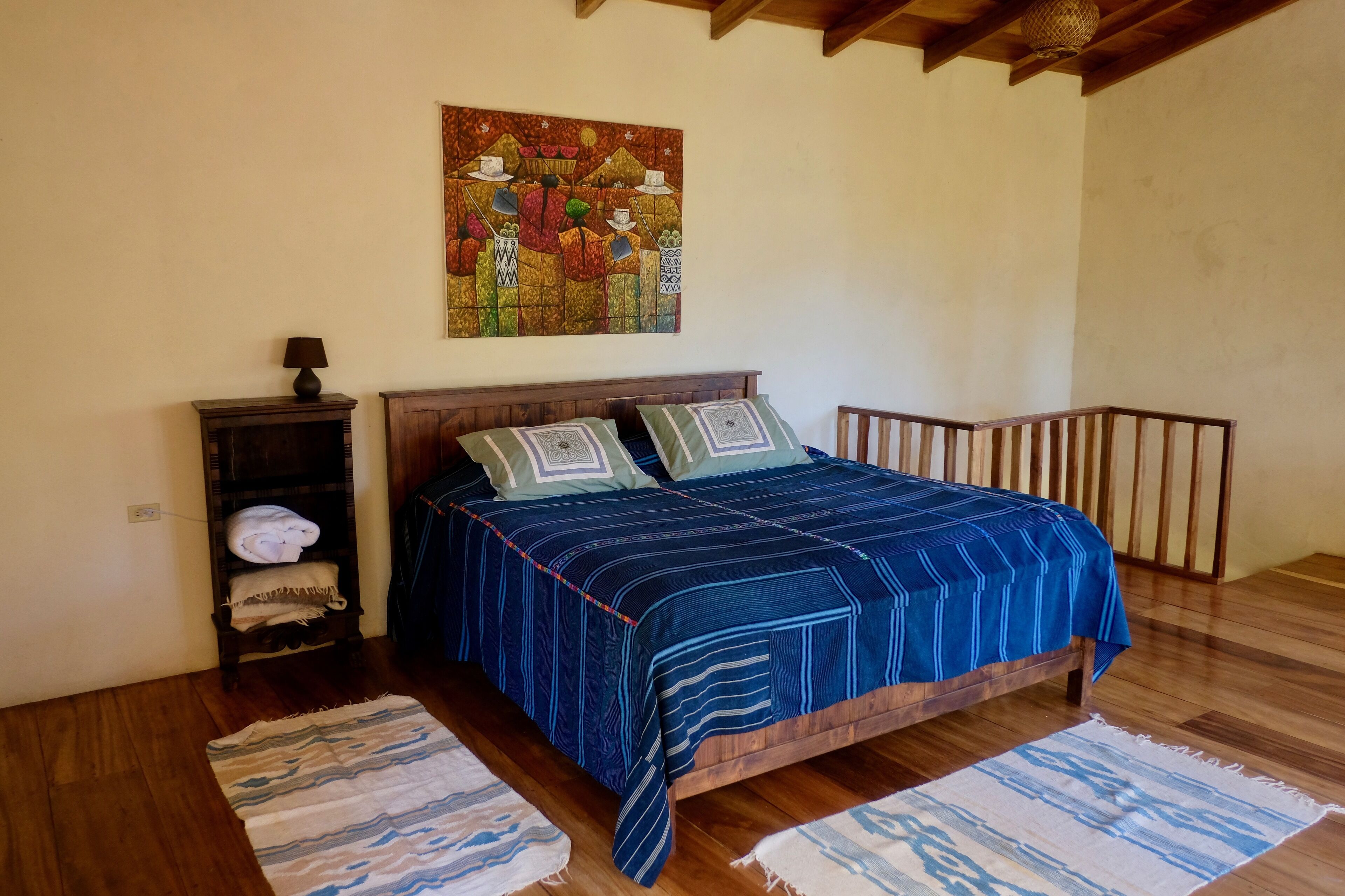 A bedroom with a wooden bed, two pillows, a nightstand, a lamp, and a painting on the wall.