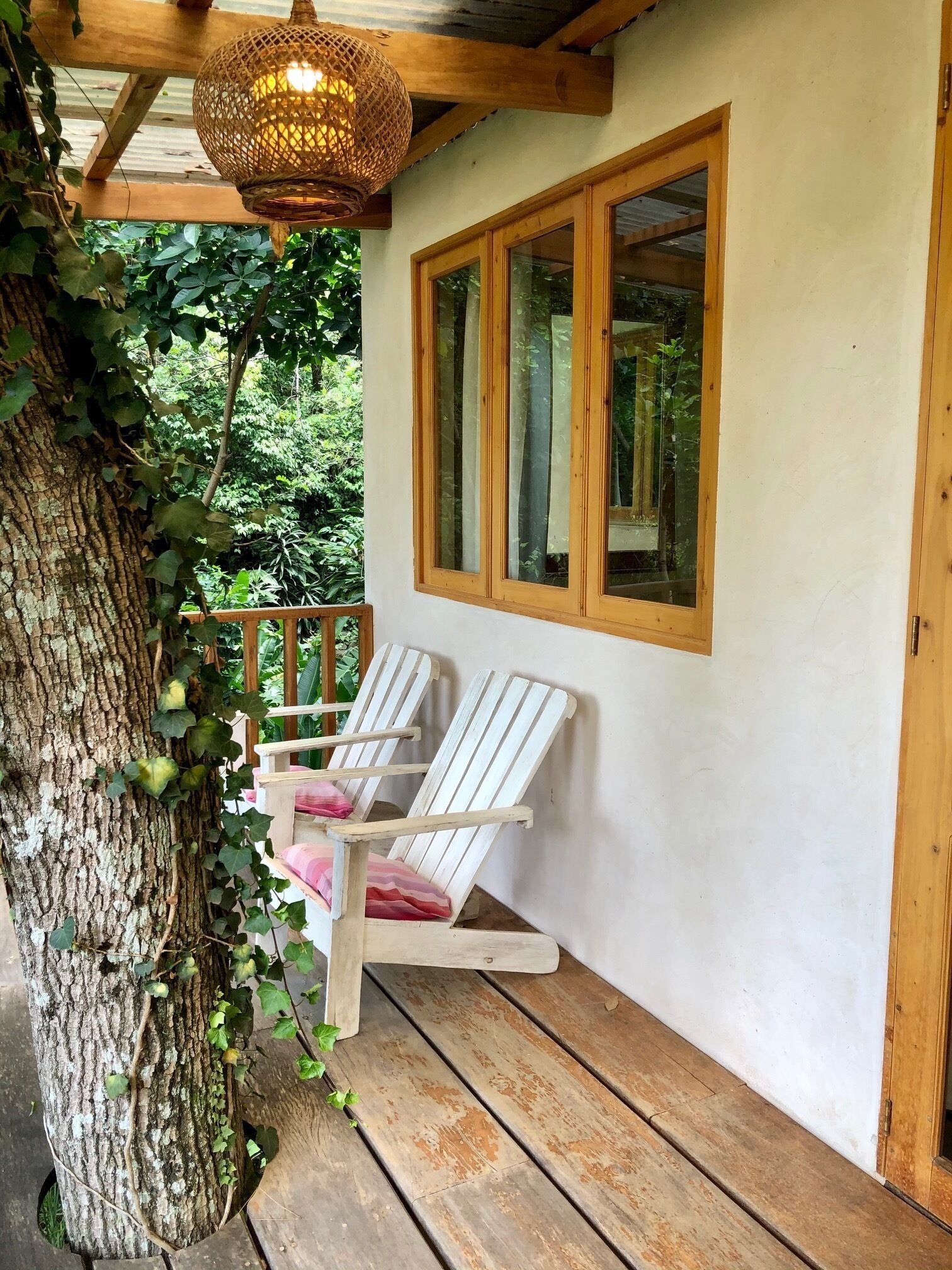 Egy fából készült verandán két fehér, Adirondack-stílusú szék, egy fonott, lógó lámpatest és egy fa kerettel ellátott ablak található.