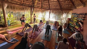 Ein Yoga-Kurs in einem Raum mit Strohdach und großen Fenstern, Holzböden und Yogamatten.