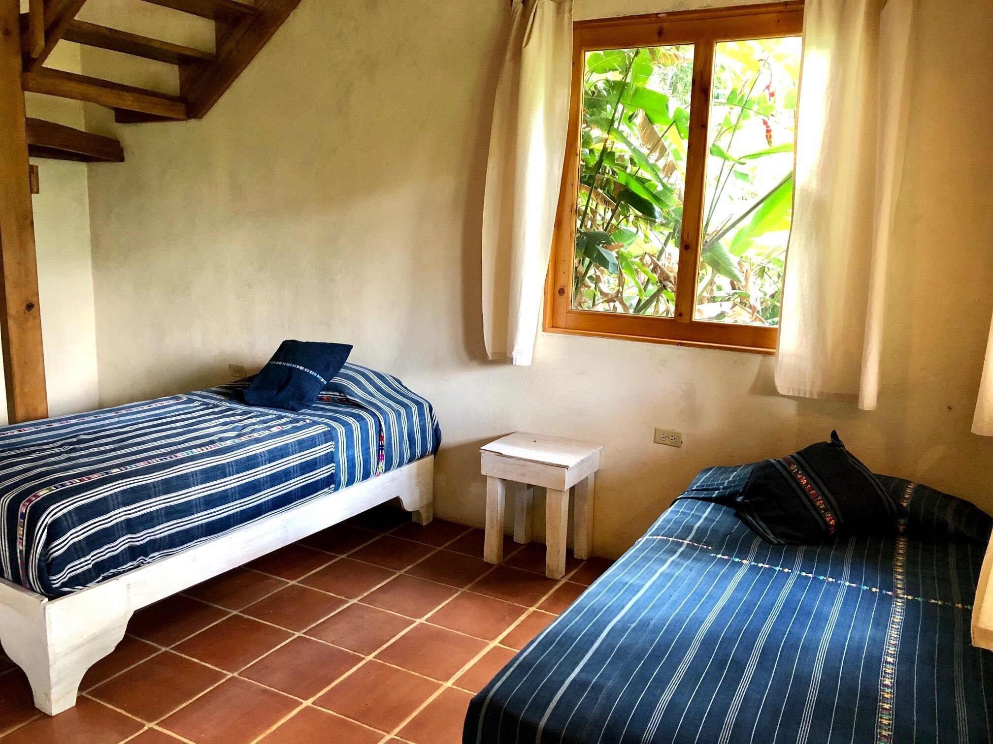 Um quarto pequeno e acolhedor, com duas camas, uma mesinha e uma janela com cortinas.