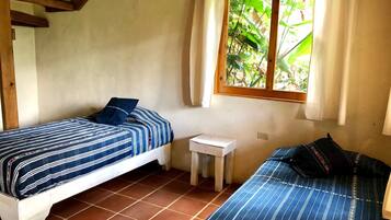 Um quarto pequeno e acolhedor, com duas camas, uma mesinha e uma janela com cortinas.