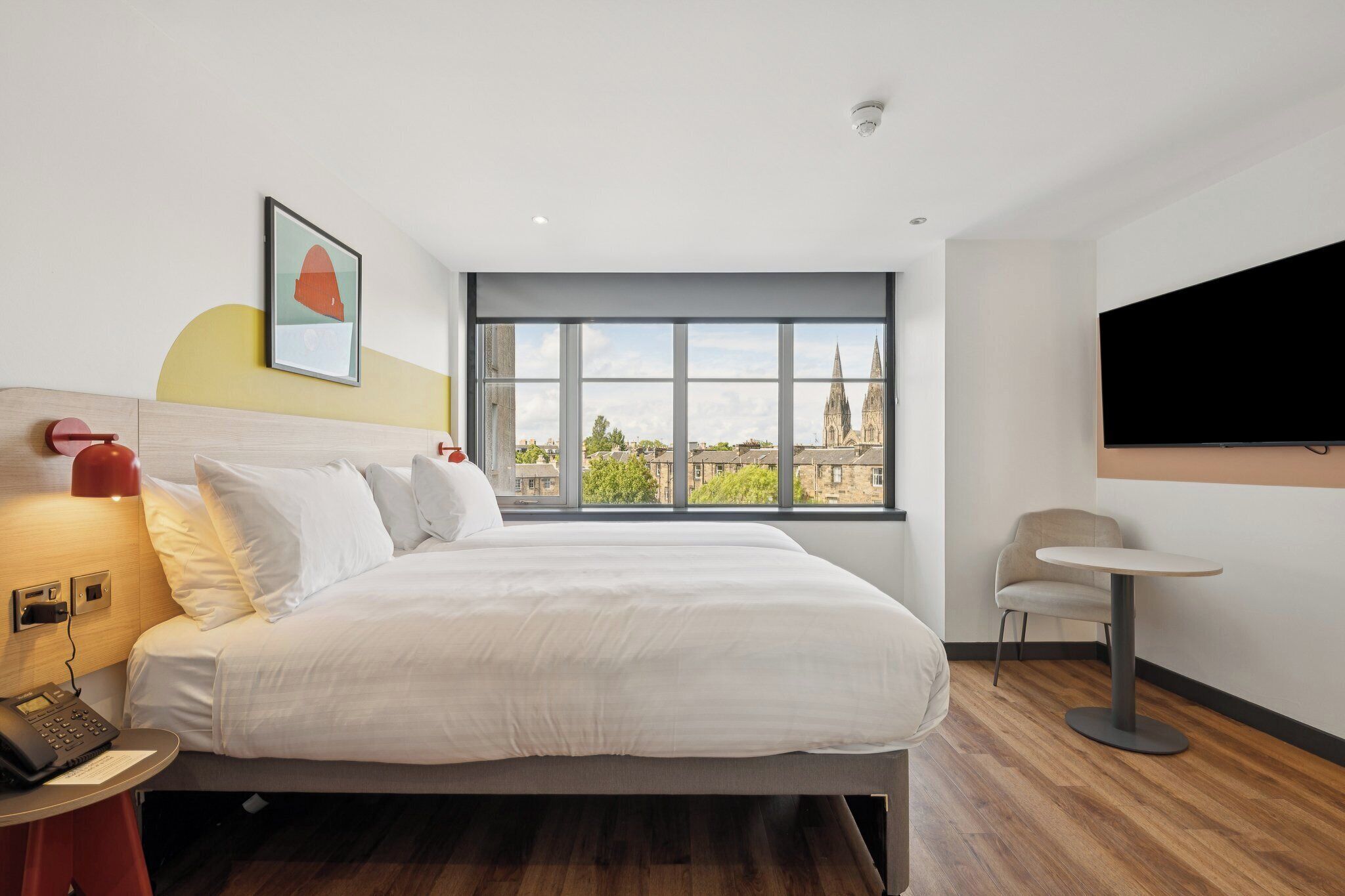 A modern hotel room with a large bed, a bedside table with a lamp, a chair, a round table, and a TV mounted on the wall.