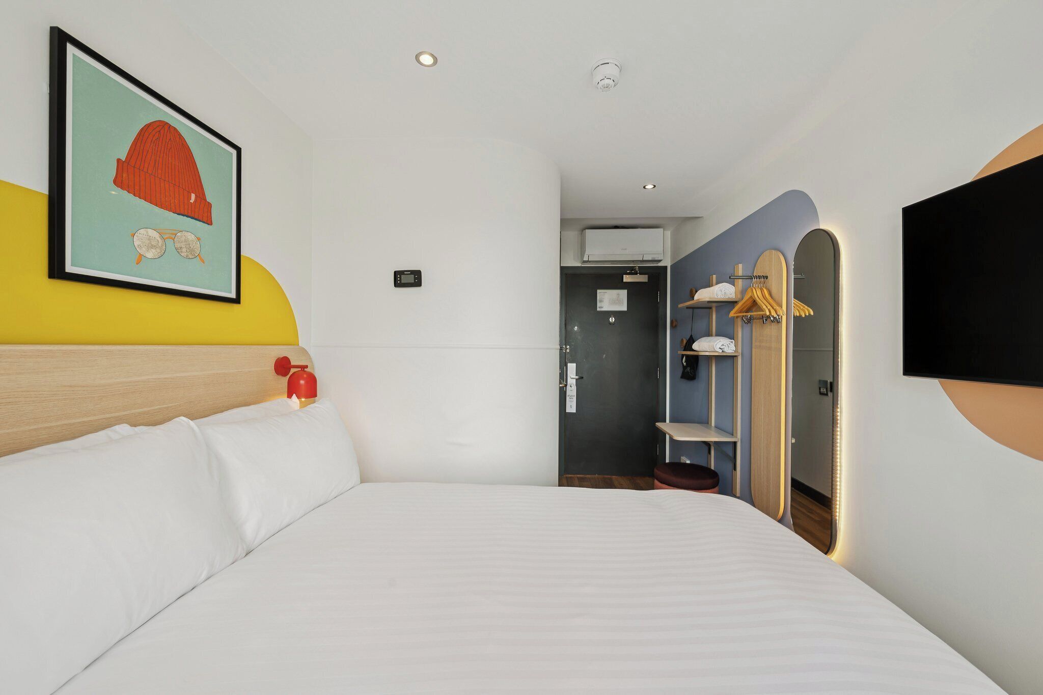 A hotel room with a bed, a wall-mounted TV, a door leading to a bathroom, and a coat rack with hangers.