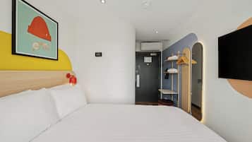 Habitación de hotel con cama, televisor montado en la pared, puerta que da a un baño y perchero con perchas.