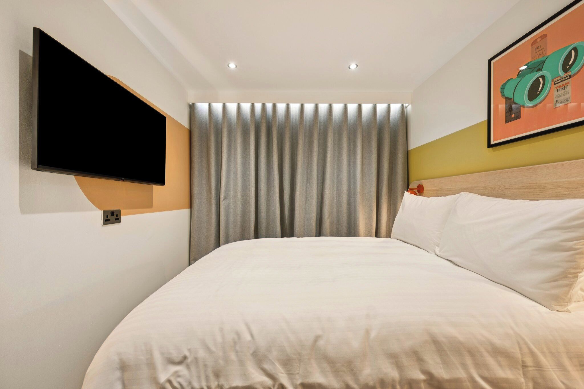 Ein Hotelzimmer mit Bett, einem an der Wand montierten Fernseher, einem Vorhang und einem gerahmten Bild.