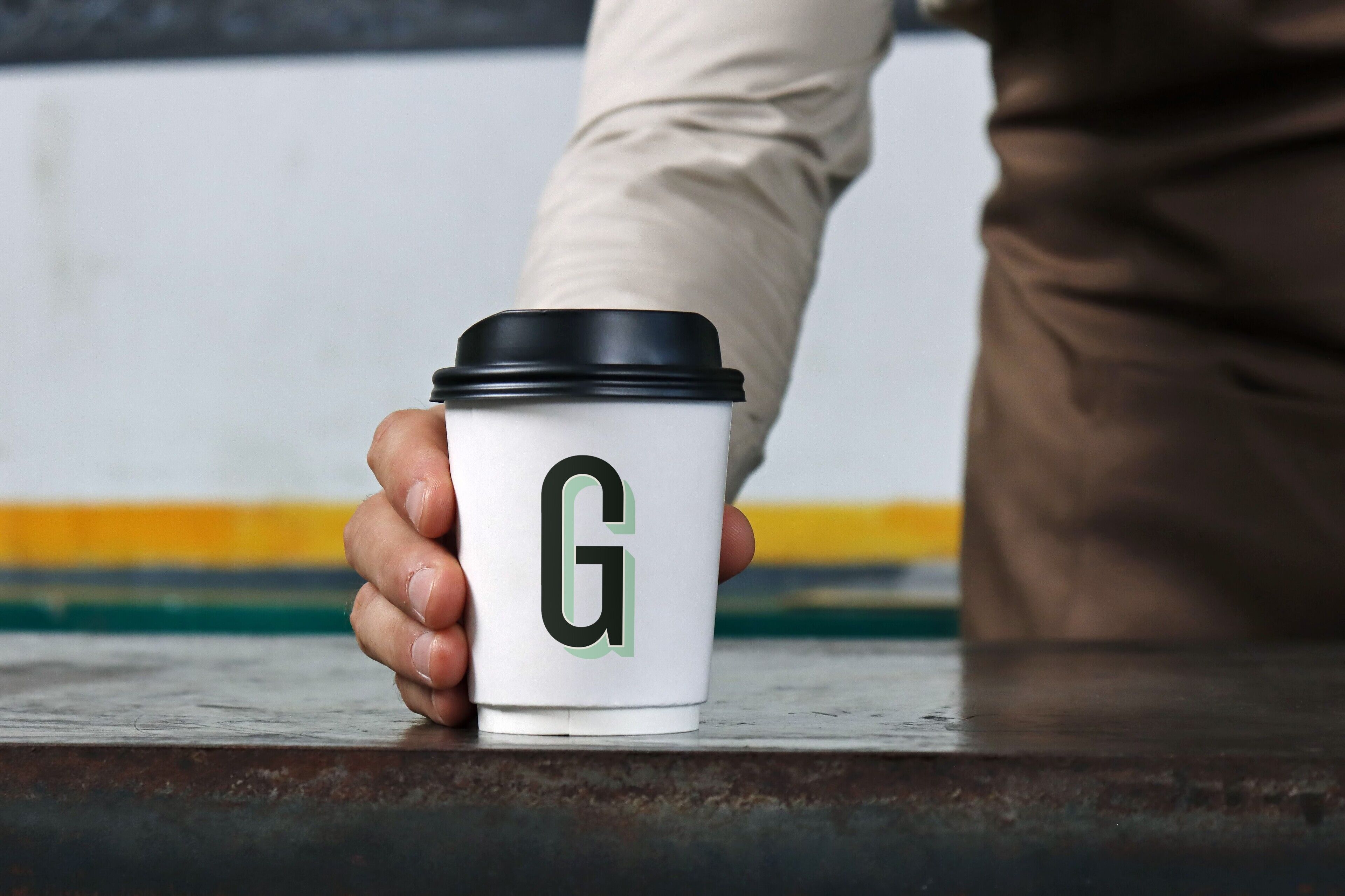 A person holding a white coffee cup with a black lid and a green letter 'G' on it.